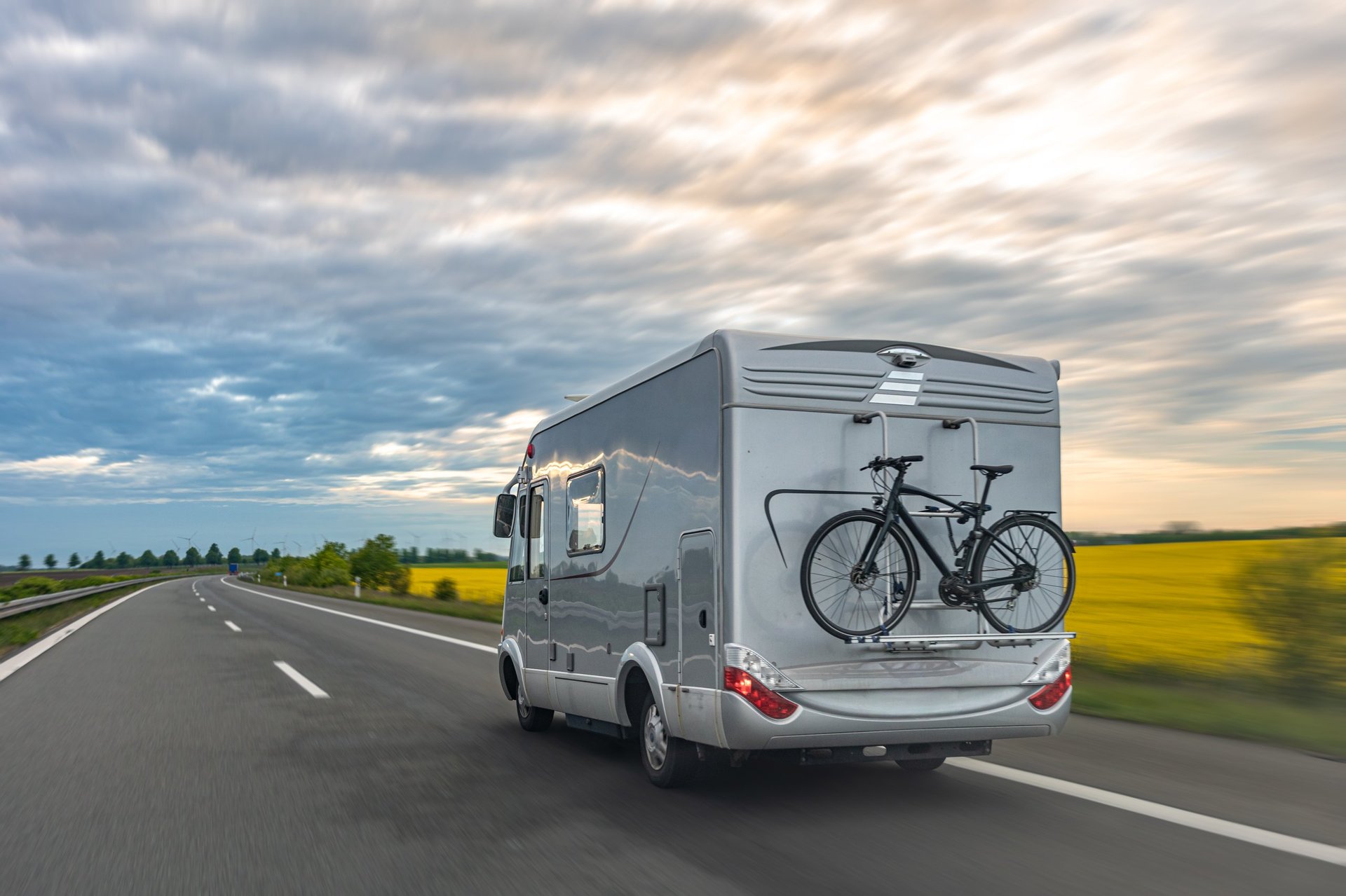 A gray motorhome drives on a highway with a bicycle mounted on the rear rack and open fields nearby