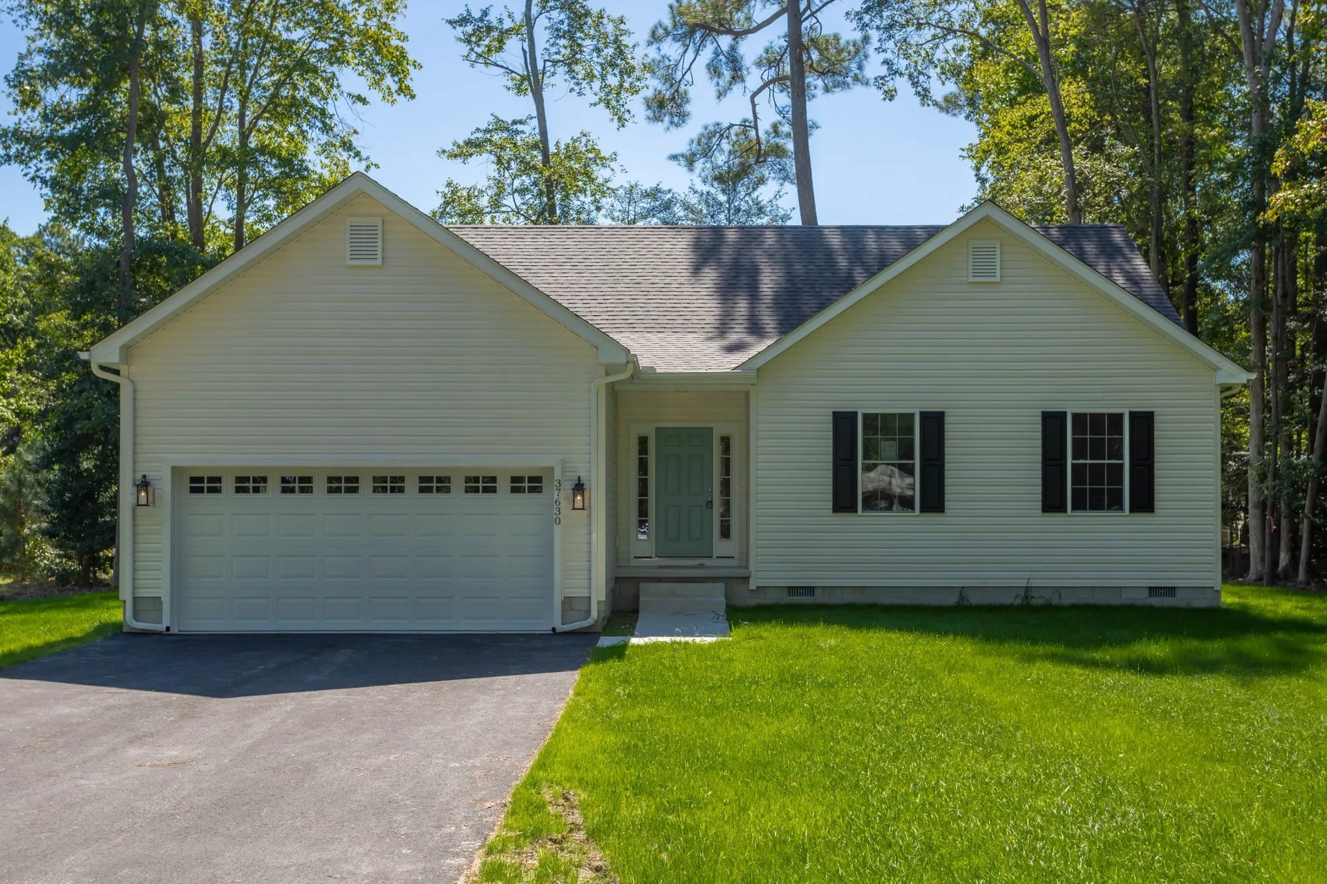 Single-story beige house with two-car garage, black shutters, and manicured lawn surrounded by tall trees