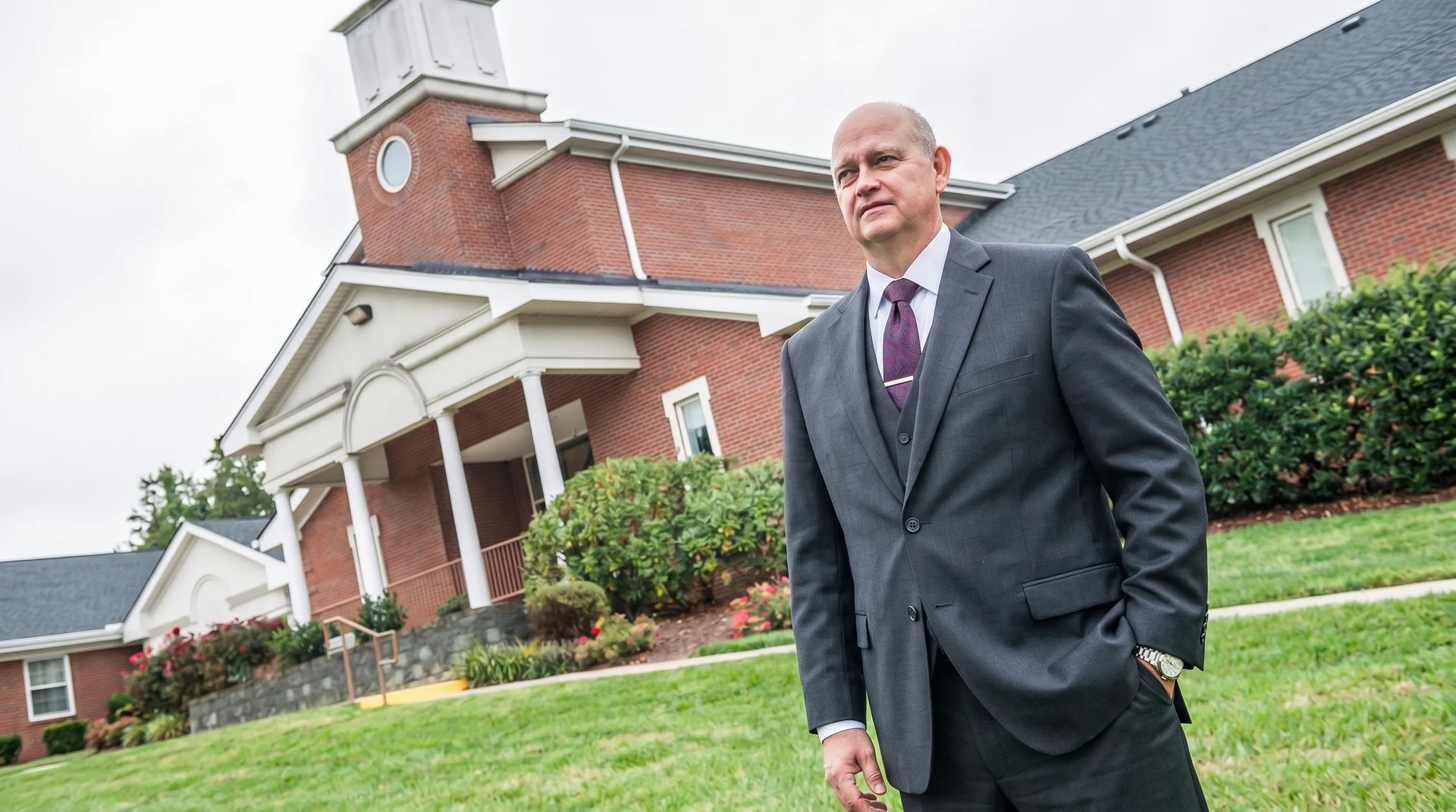 Bald man in dark suit and purple tie standing on grass in front of brick church building with steeple and white columns