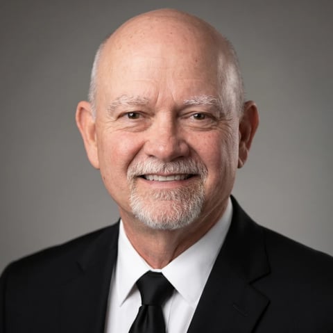 Bald man with gray beard in professional black suit and black tie, smiling at camera against gray background
